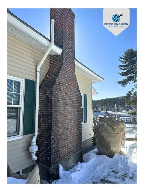 A house with pastel yellow siding, green shutters, and a brick chimney with white piping against a snowy landscape and blue sky.