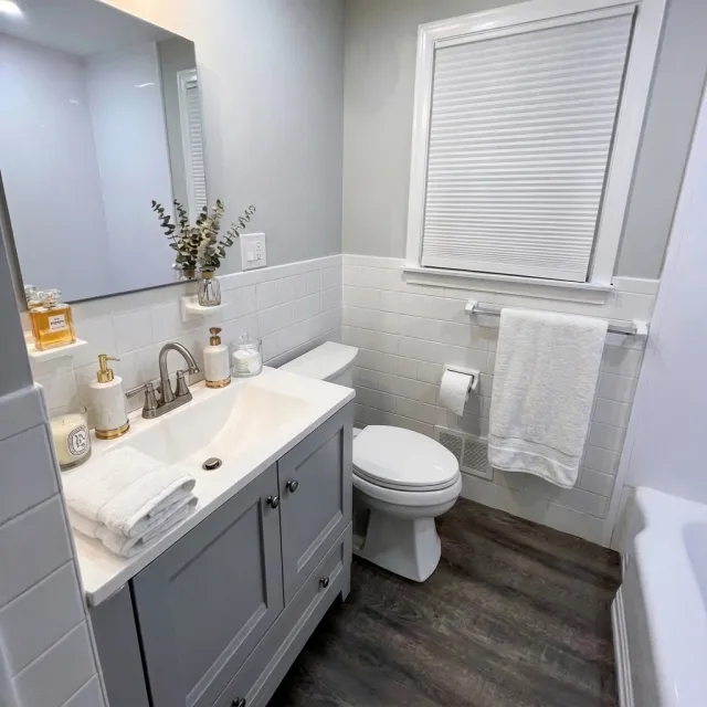 A small bathroom with a light gray vanity, white countertop, and a stainless steel faucet. The sink area has soap dispensers and a small towel on top. There is a mirror above the vanity. The toilet is next to a wall with white subway tiles half-way up, and a window with a closed blind above it. A white towel hangs on a bar below the window. The bathroom floor is dark wood.