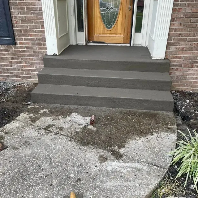 Front steps of a brick house with a wooden door, concrete walkway, and small landscaping bed.