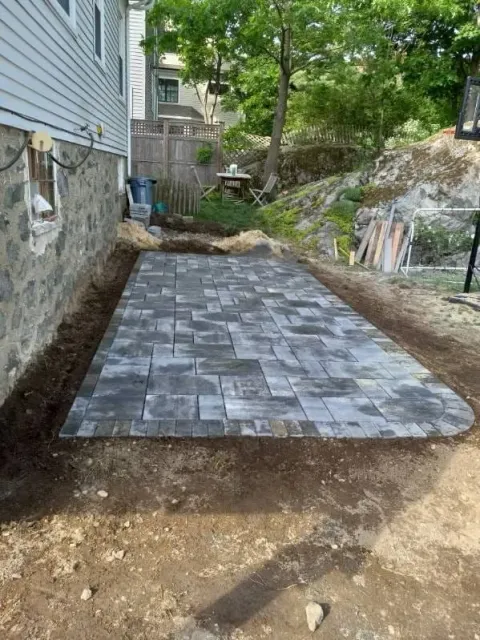 Stone paver patio under construction next to a house and hillside. Earthy tones and gray pavers.