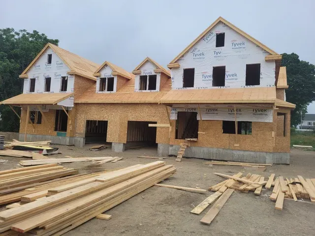Partially built two-story house with exposed wood framing and lumber piles in front