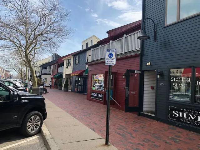 A brick-paved street lined with colorful shops and parked cars. A handicap parking sign is centered in the scene.