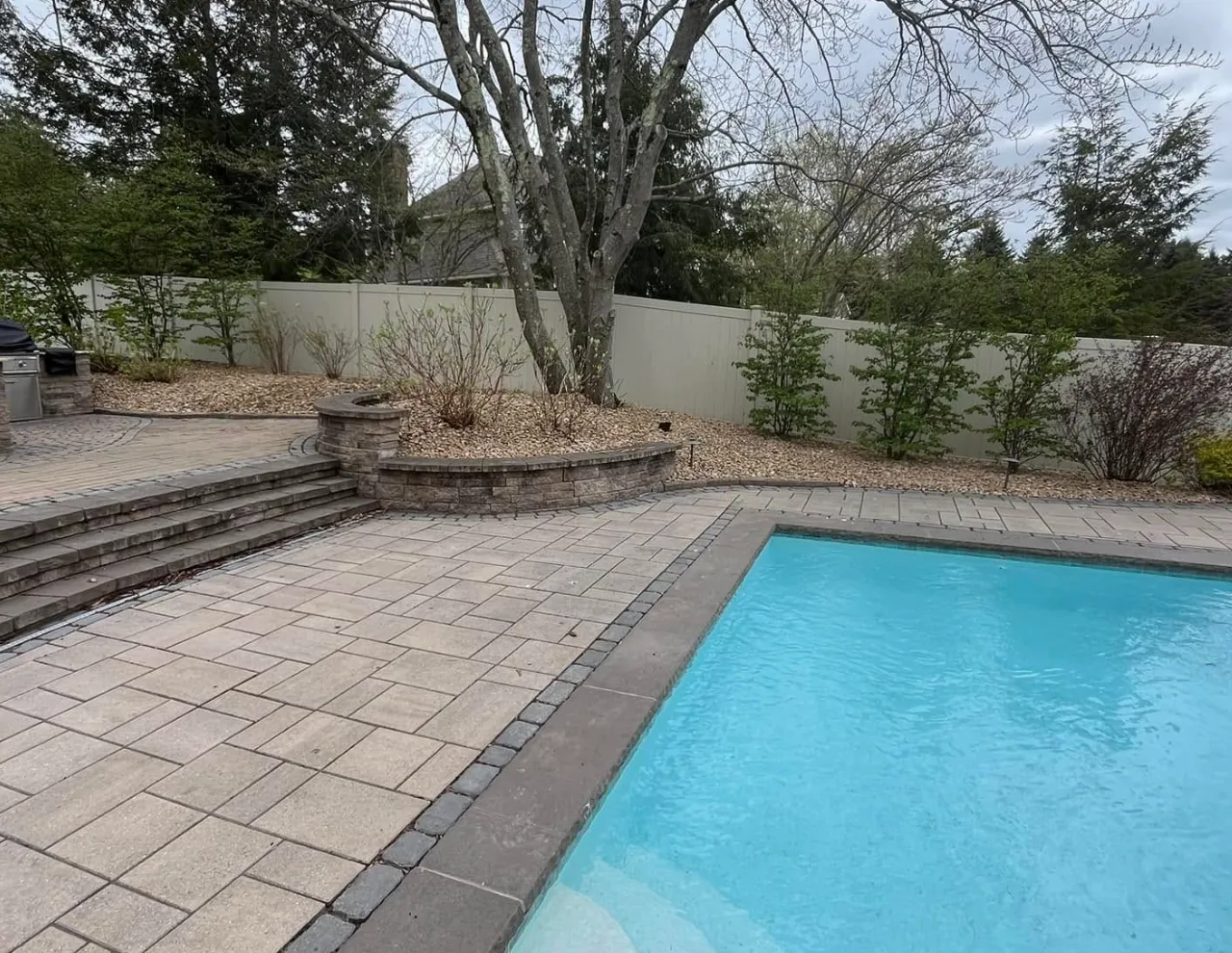 Backyard with swimming pool, stone patio, steps, trees, and a white fence.