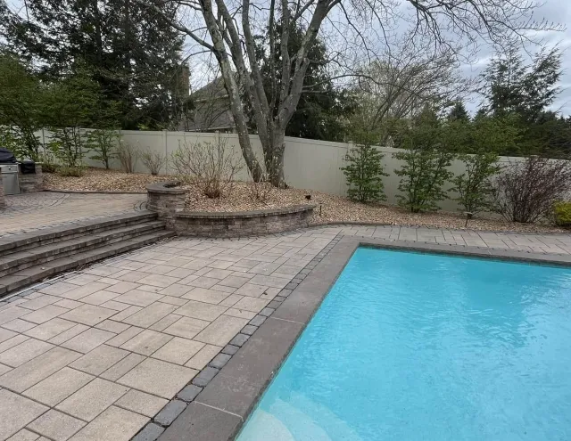 Backyard with swimming pool, stone patio, steps, trees, and a white fence.