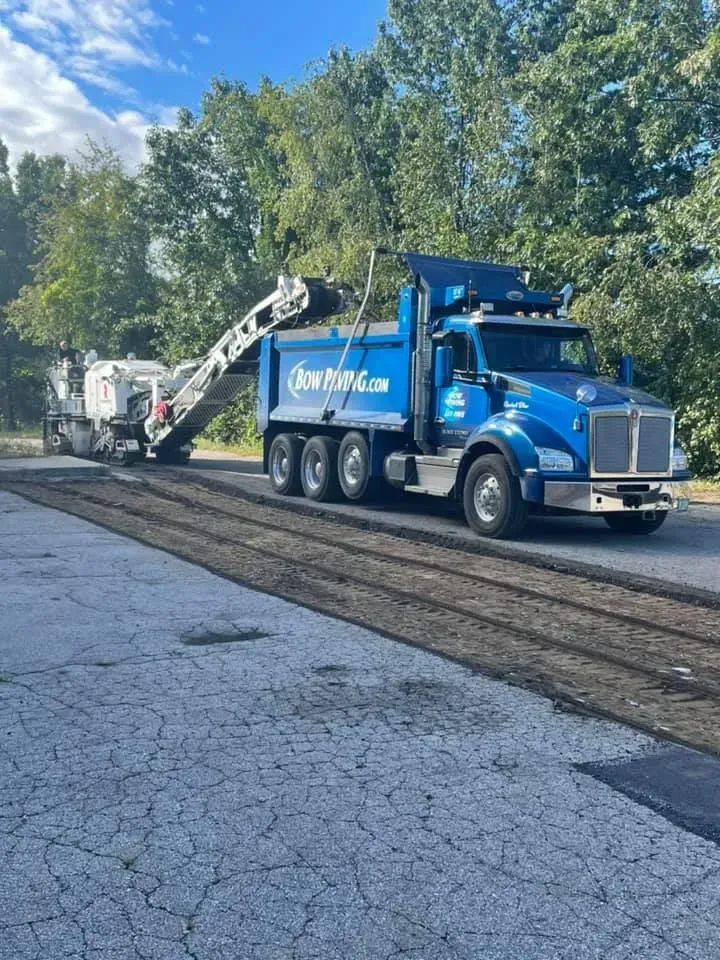 A blue dump truck is parked on an asphalt road next to a white milling machine during a road repair project.