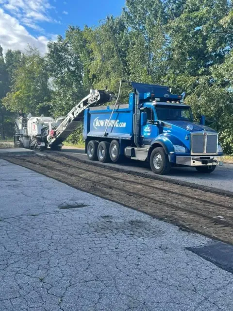 A blue dump truck is parked on an asphalt road next to a white milling machine during a road repair project.