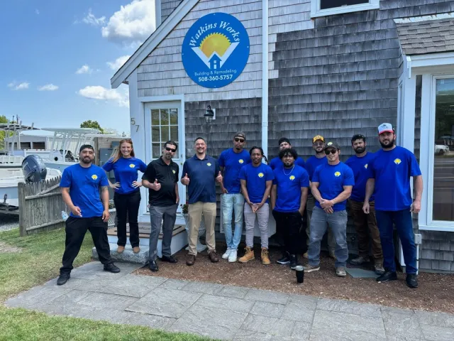 Group of twelve workers standing outside a building with a sign reading 'Watkins Works Building & Remodeling.' Cape Cod contractor providing remodeling and renovation services.