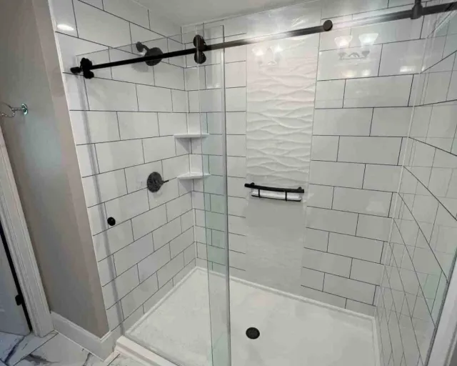 A newly remodeled walk-in shower in a South Shore Boston home featuring large subway-style wall panels, matte black hardware, recessed shelving, and a textured white accent wall. The frameless glass doors and marble-look tile flooring highlight Bay State Bath’s clean, high-end installation style.