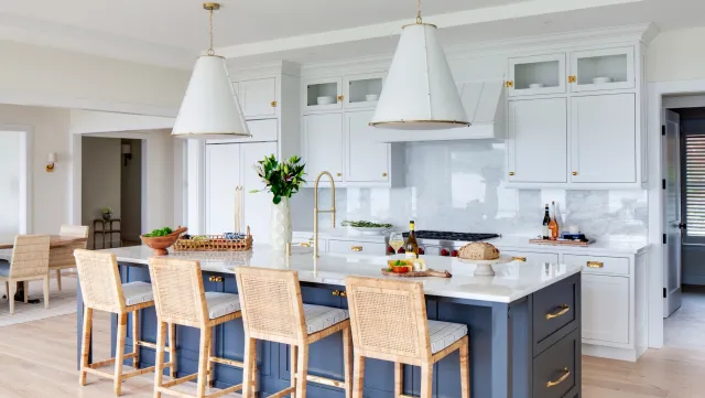 Modern white kitchen with a central island, blue base, white marble countertop, and six barstools with rattan backs. The kitchen has white cabinets with glass doors, gold handles, and a marble backsplash. Two large white pendant lights hang above the island. Kitchen counter has a wine bottle, glasses, and a loaf of bread.
