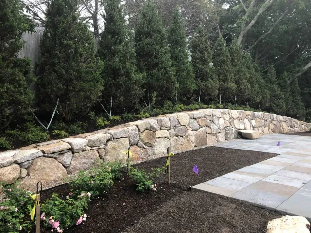 A stone retaining wall curves along a patio with a garden bed in the foreground and tall evergreen trees in the background.