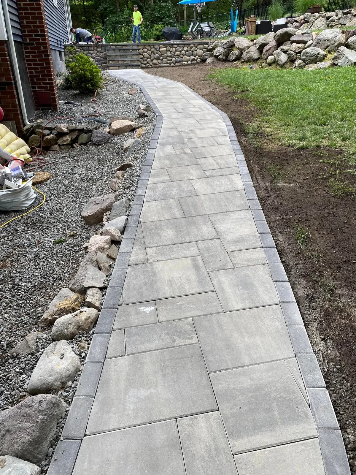 A stone walkway curves through a yard. Gray pavers are bordered by darker stones and gravel.