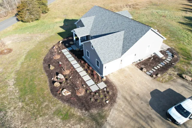 Aerial view of a house in a semi-rural area with a gray roof, white exterior walls, and a small front porch. The yard has a landscaped garden with stone pathways and a few trees, surrounded by grassy fields and a road.