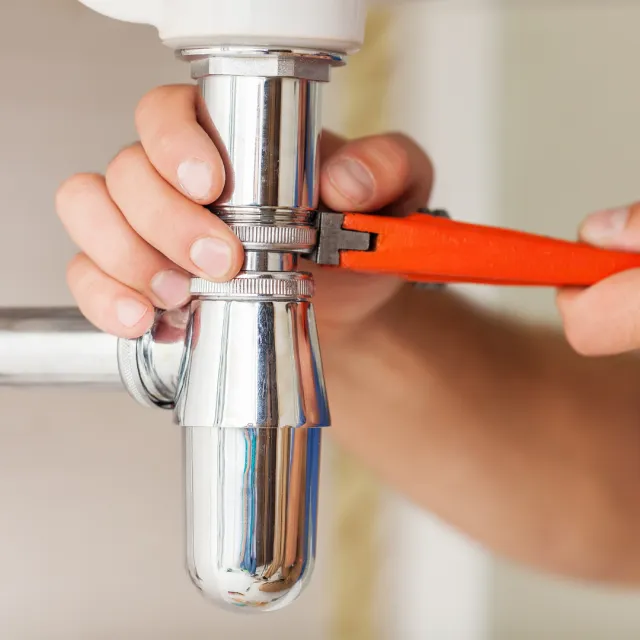Person using a wrench to tighten or loosen a shiny metal pipe under a sink.