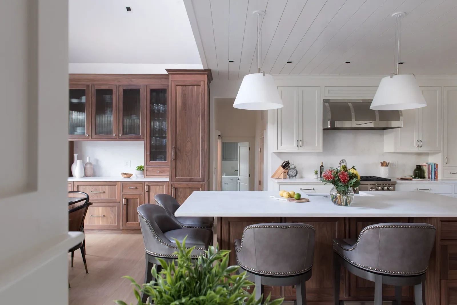 Modern kitchen with white cabinets, a large white marble island, a wooden wall of cabinets, and a stainless steel stove with a range hood. Two white pendant lights hang over the island, which has a vase of flowers and some fruit on it. Barstools are positioned around the island, and there is a plant in the foreground.