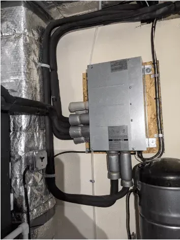 A silver HVAC system stands prominently, featuring insulated ducts, a mounted control panel, and visible pipes and wiring against the wall. Explore flexible finance options to make this essential upgrade more accessible.