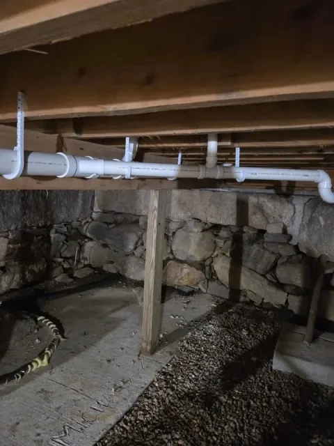 Underneath a wooden floor with plumbing pipes running along the joists, supported by vertical wooden posts, with a stone wall in the background and gravel ground below.