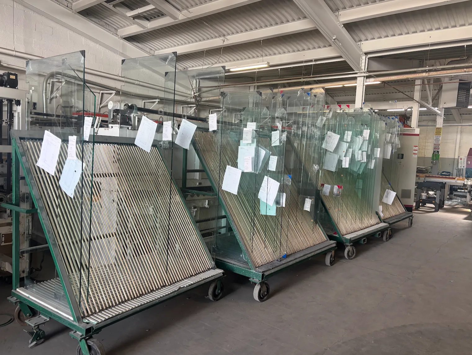 Rows of glass sheets stored on trolleys in a manufacturing or warehouse setting, with each sheet labeled with papers attached.