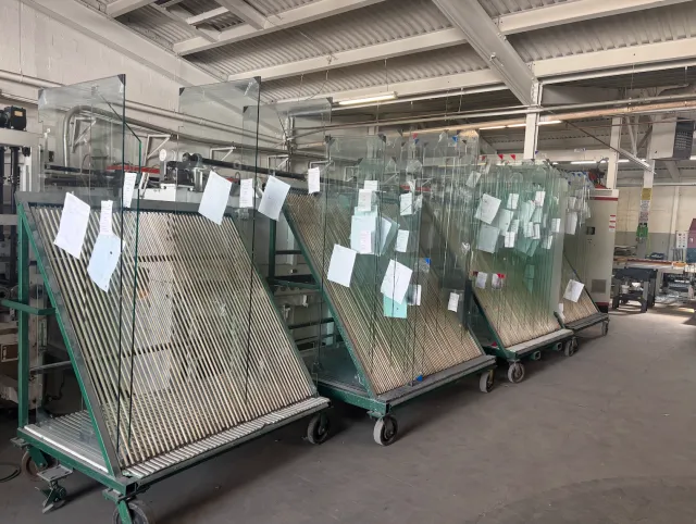 Rows of glass sheets stored on trolleys in a manufacturing or warehouse setting, with each sheet labeled with papers attached.