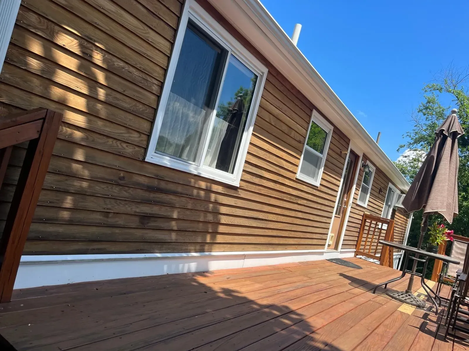 Wooden house deck with brown siding, white-trimmed windows, and patio furniture under a blue sky
