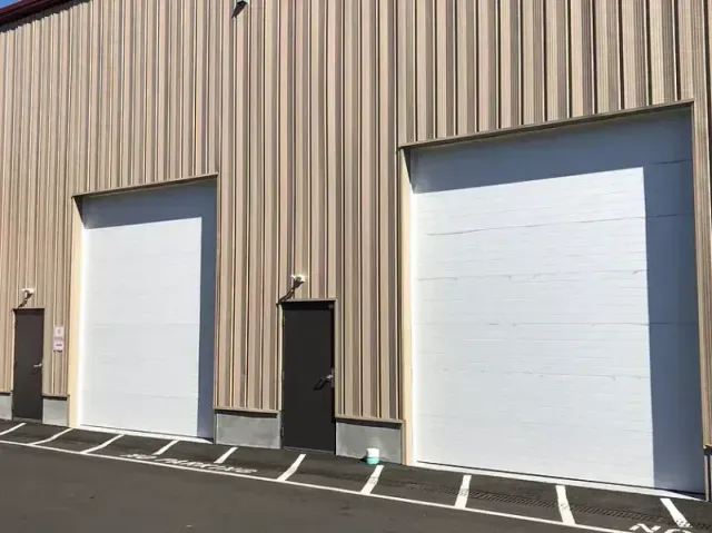 Exterior view of a beige industrial building with two large white loading doors and two dark side doors.
