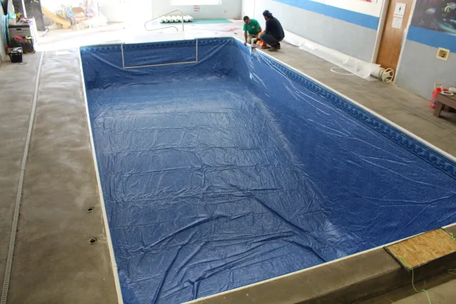 An indoor swimming pool under renovation, with a blue tarp lining the empty pool, workers in green and blue shirts working nearby, construction supplies, and tools around the pool area.