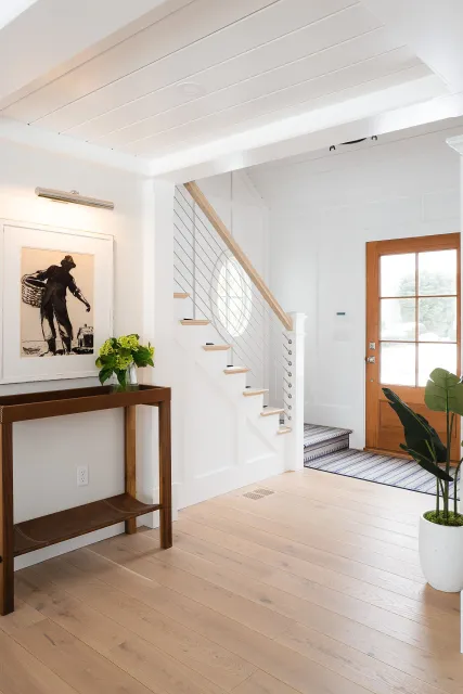 Hardwood floor installation in a Scituate entryway featuring white walls, a wooden front door, a staircase with a wood handrail and white railing, framed artwork, a console table with a green plant, and a striped rug.