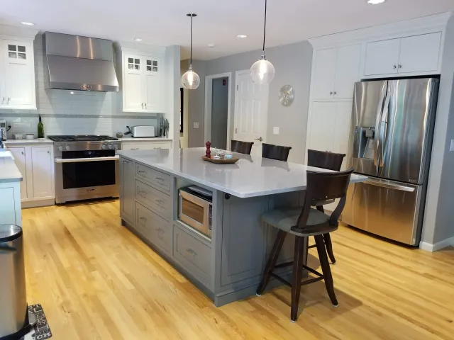 Modern kitchen with white cabinets, stainless steel appliances including a refrigerator and oven, a kitchen island with drawers, bar stools, and hanging pendant lights.