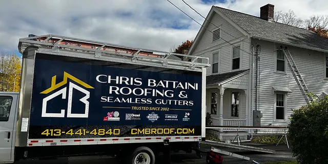 Chris Battaini Roofing - A roofing company truck parked in front of a white house, with ladders set up for roof work. The truck advertises Chris Battaini Roofing & Seamless Gutters.
