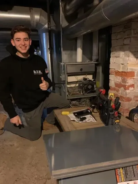 HVAC technician kneels near furnace, giving thumbs up. Smiling, black sweatshirt, tools, basement setting.
