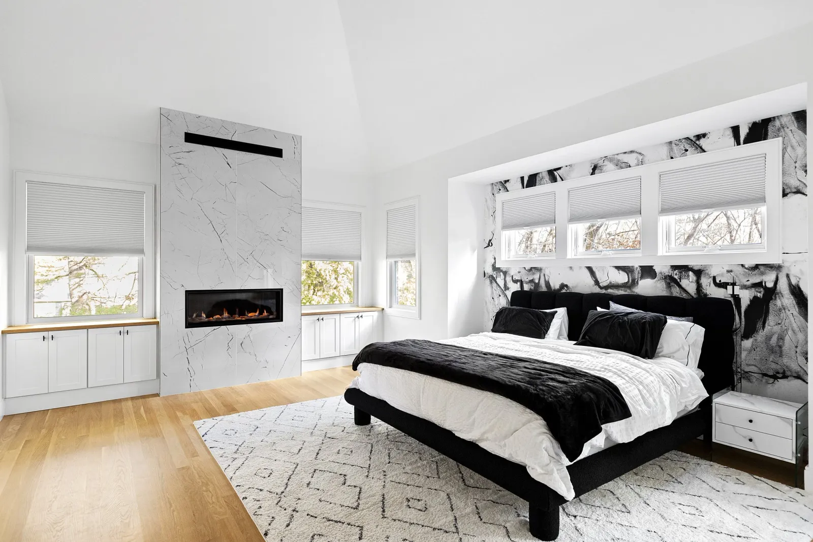 Modern bedroom with black and white color scheme, featuring a bed with black upholstery, white linens, and black pillows, a white patterned rug, windows with blinds, a black and white abstract feature wall, and a wall-mounted fireplace.