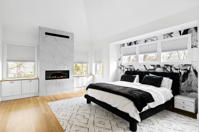 Modern bedroom with black and white color scheme, featuring a bed with black upholstery, white linens, and black pillows, a white patterned rug, windows with blinds, a black and white abstract feature wall, and a wall-mounted fireplace.