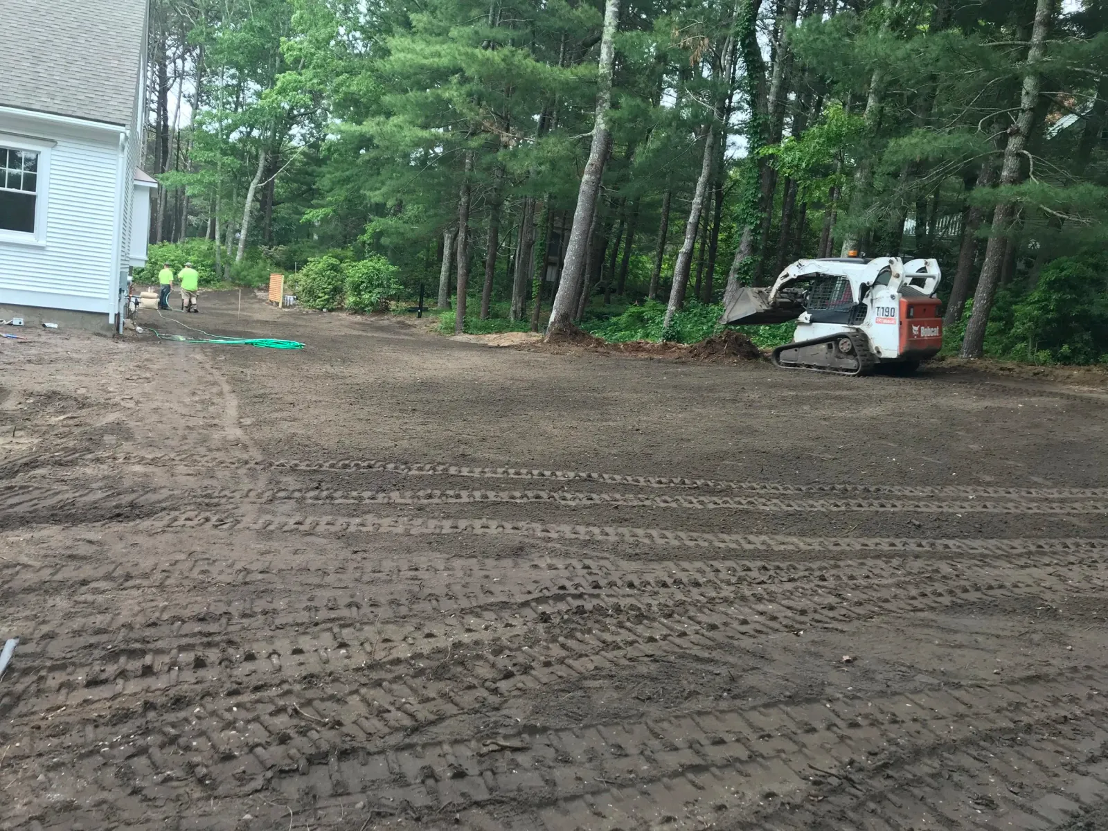 Construction site with a white house, two workers, a Bobcat T190 skid steer, and a cleared dirt area surrounded by trees.