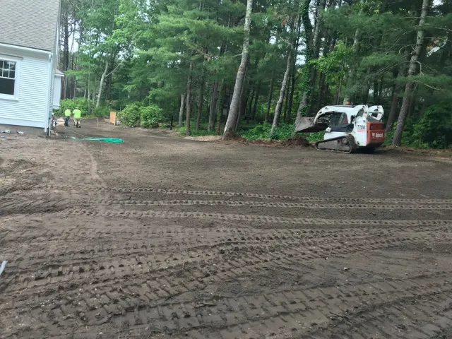 Construction site with a white house, two workers, a Bobcat T190 skid steer, and a cleared dirt area surrounded by trees.