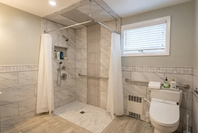 Bathroom with walk-in shower, white curtains, and a toilet with a bidet function. The shower has marble tiles, a recessed shelf with toiletries, and a handheld showerhead. There is a small window with blinds, a tiled wall border, and a heater below the window. Toiletries are placed on top of the toilet tank.