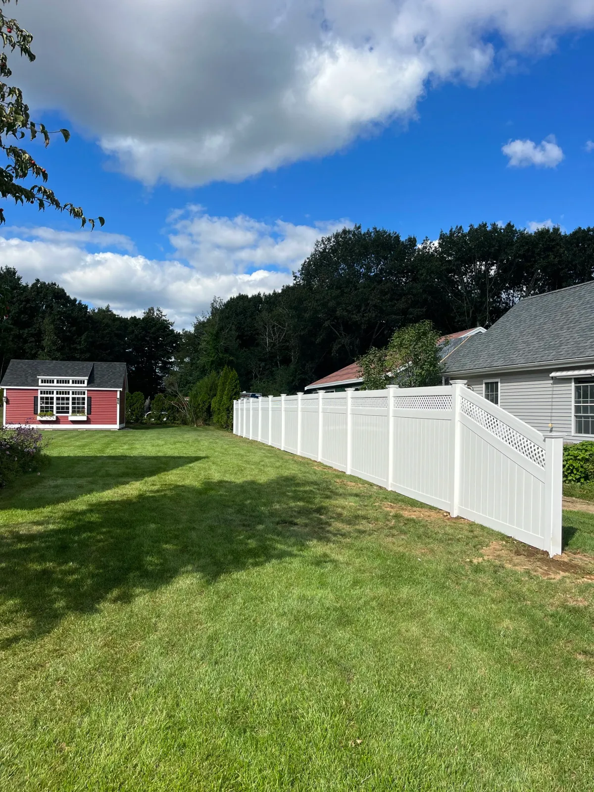 A backyard with green grass, a white privacy fence, a gray house with a window and a porch on the right, and a small red and white shed with a window on the left. There are trees and bushes in the background, and a partly cloudy blue sky overhead.