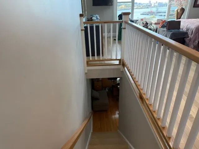 Staircase leading downwards. White walls and railing, wooden handrail, view to lower floor with furniture.