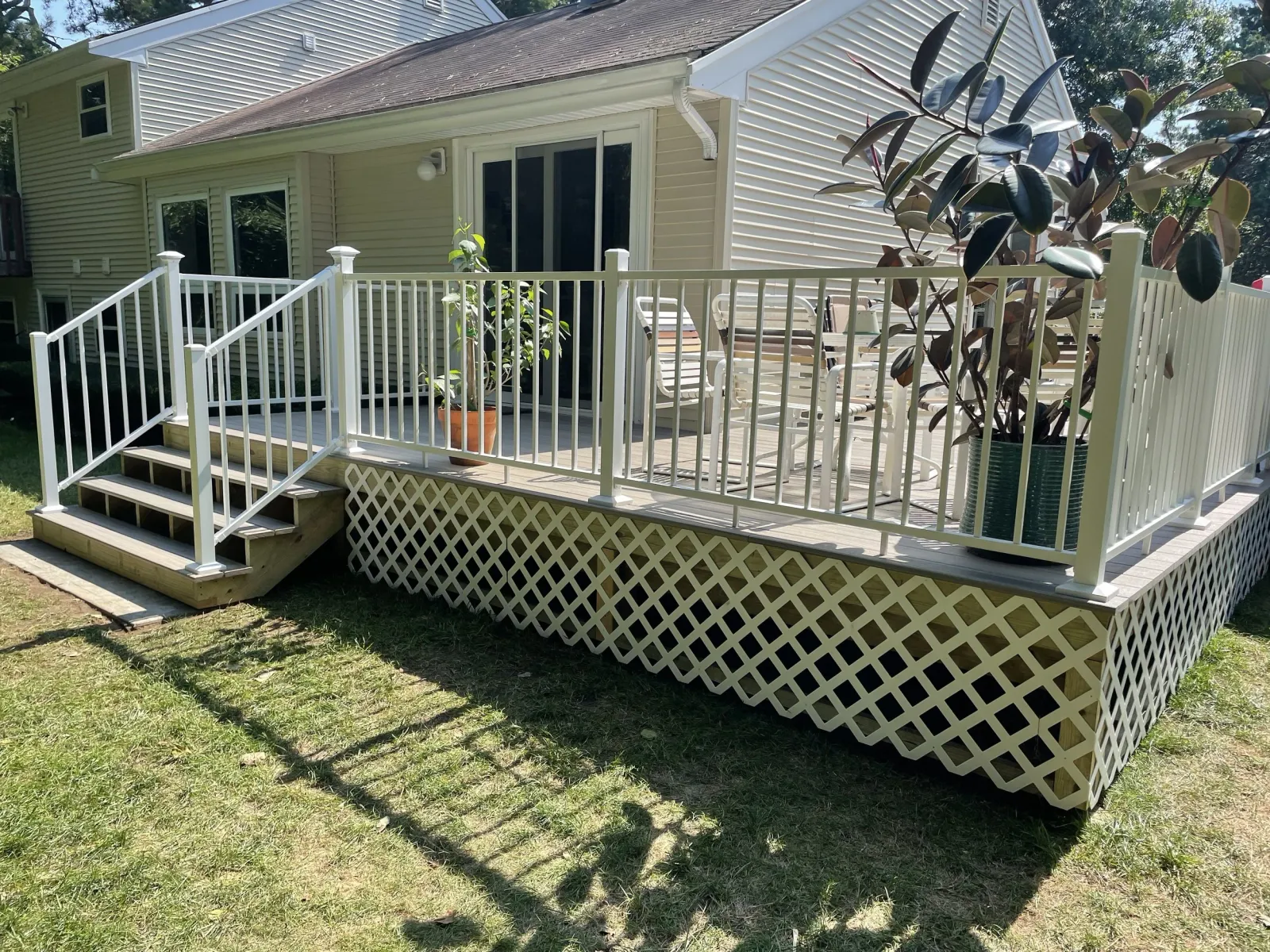 Backyard deck with white railing, stairs, potted plants, chairs, and a lattice skirting under a beige house