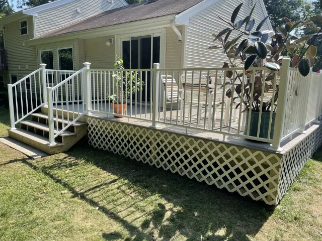 Backyard deck with white railing, stairs, potted plants, chairs, and a lattice skirting under a beige house