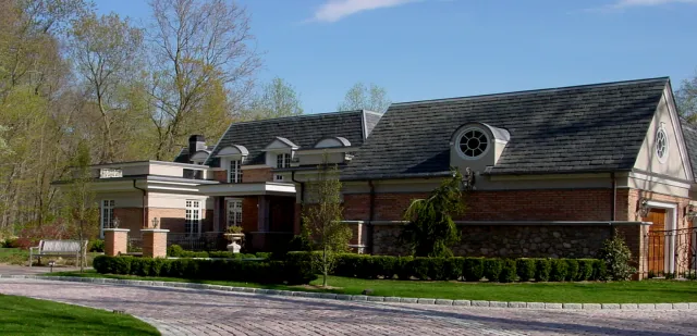 Large brick house with multiple roof levels, arched dormer windows, circular window accents, stone and brick exterior, and landscaped front yard with a cobblestone driveway.