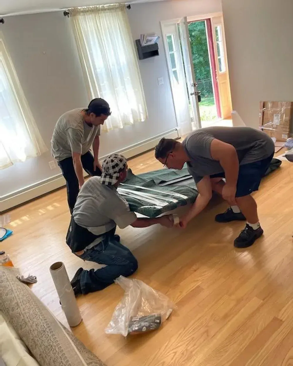 Three people moving a green tarp-covered object inside a room with hardwood floors and bright windows.