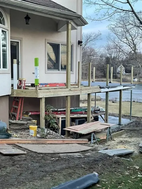 Deck construction underway, house exterior. Wooden frame, supports, tools, and materials visible. Overcast day.