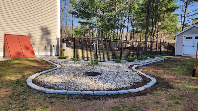 A rock garden bordered by gray stones in a yard, with a small shed and tall trees in the background.