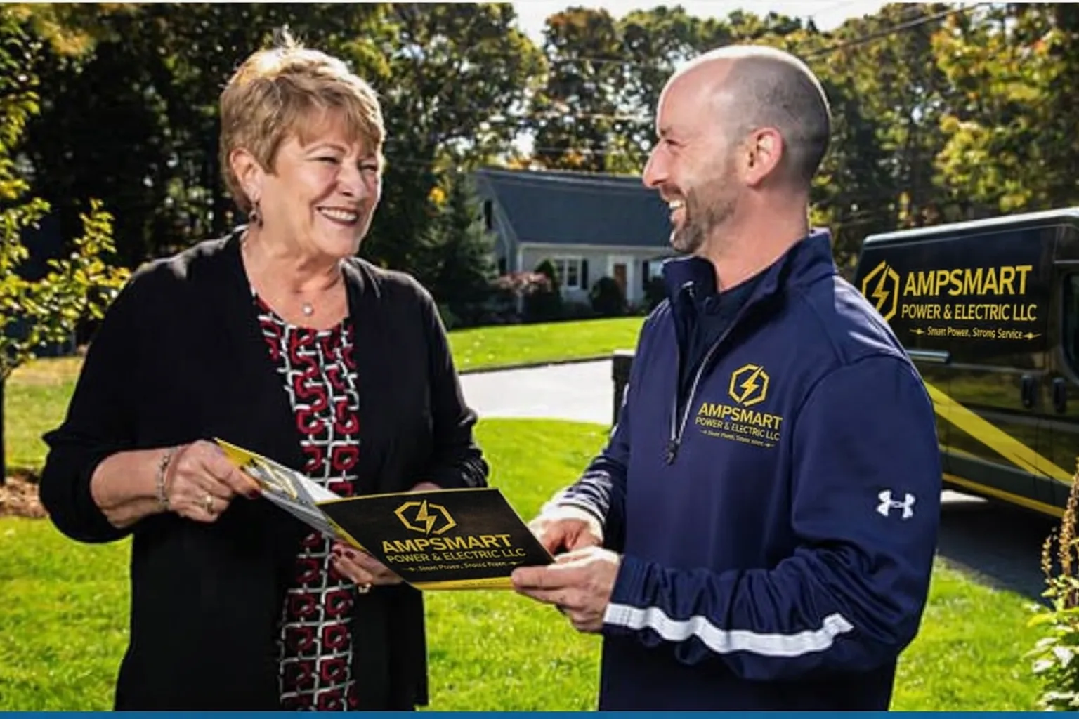A woman and a man shake hands outdoors, exchanging a brochure for Ampsmart Power & Electric LLC. The woman smiles and wears a black jacket over a red patterned top. The man, in a blue athletic jacket with Ampsmart logo, smiles back. There's a van with the same logo in the background on a sunny day.