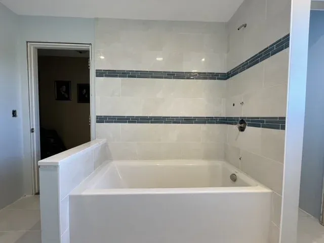 Bathroom with a white bathtub, partially tiled walls with white and blue mosaic accents, and an open doorway leading to another room.
