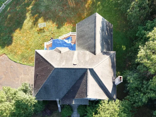 Overhead view of a house with dark gray roof, surrounded by green trees and lawn, with a blue tarp on the roof.