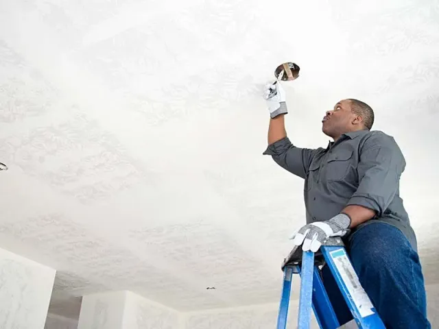 A man is sitting on a ladder fixing a light fixture on the ceiling.