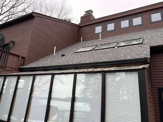 Two-story brown house with snow-dusted roof and large windows under an overcast sky