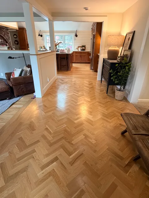 View of a living room with hardwood herringbone parquet flooring, partially visible furniture including a leather armchair, a black dresser with decorative items and paintings, and a kitchen in the background with wooden cabinets, a central island, and large window letting in natural light.