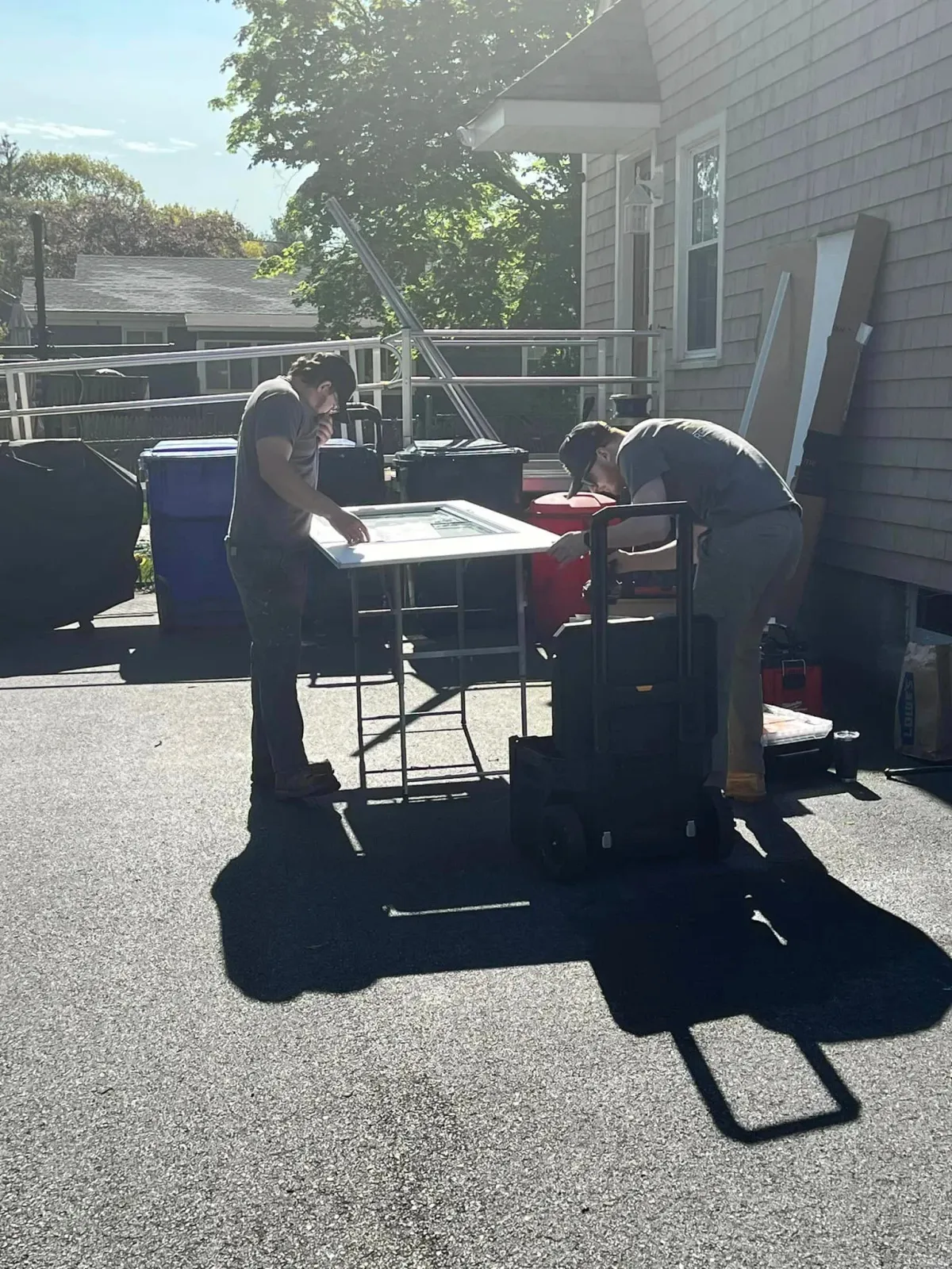 Two people working on a table outside a house on a sunny day, with shopping carts and bins nearby, and a balcony with a metal railing in the background.