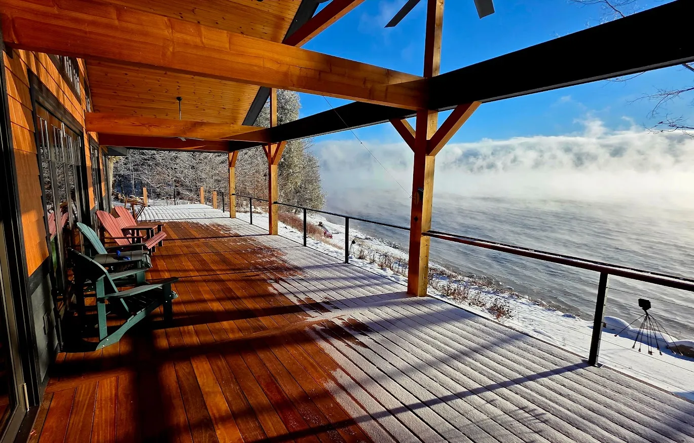 Covered waterfront porch with cumaru hardwood decking, a timber-frame ceiling, and cable railing overlooking a misty lake by Toensing Construction in Rhode Island